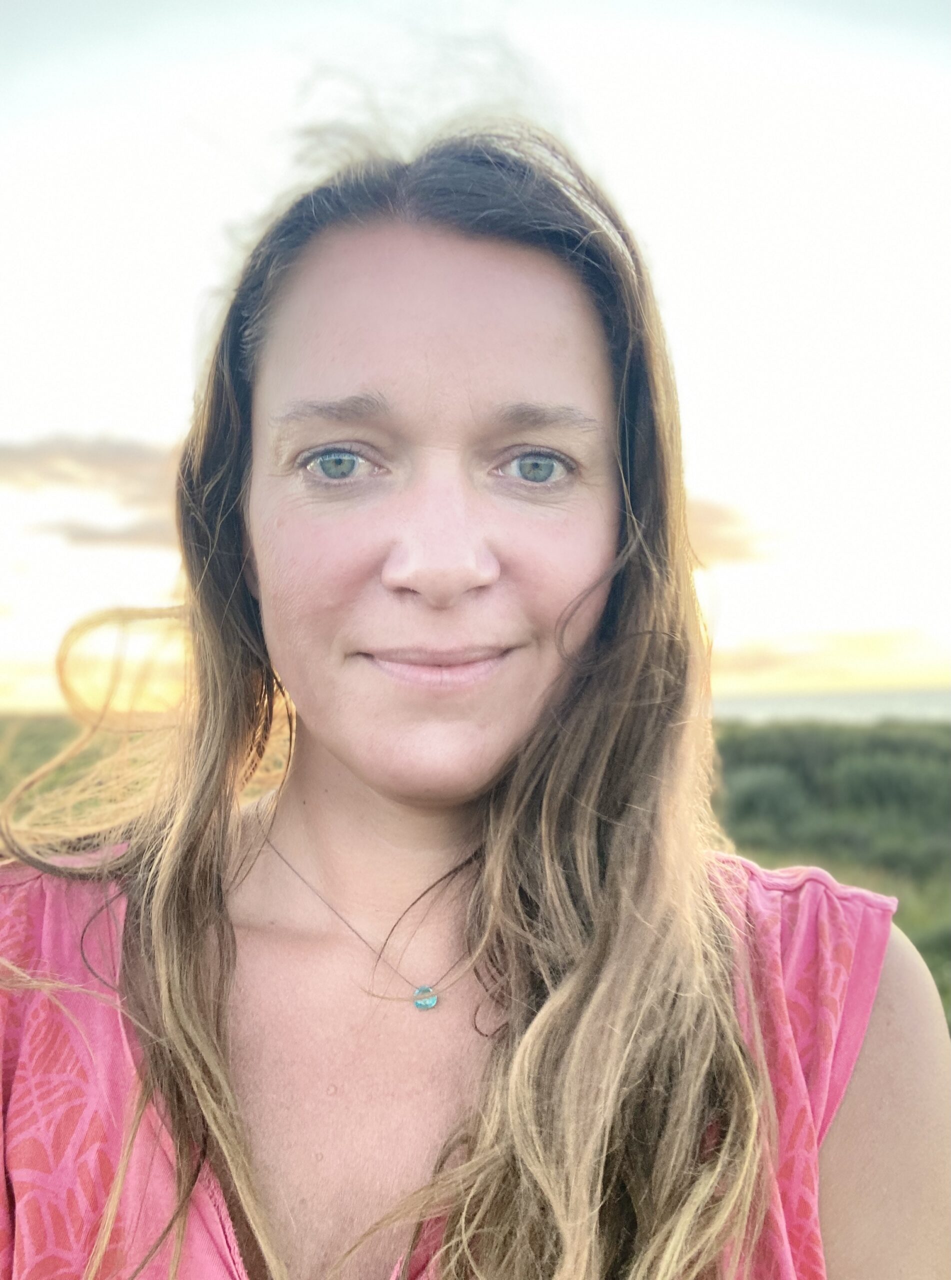 Tasja Nummert, Heilpraktikerin in Lübeck, steht vor einem Feld. Eine Person mit langen Haaren und einem rosa Oberteil steht im Freien vor einer grünen Kulisse und einem hellen Himmel. Sie hat einen ruhigen Gesichtsausdruck, trägt eine Halskette mit einem kleinen Anhänger und ihre Präsenz scheint leise zu sagen: „Über mich.“