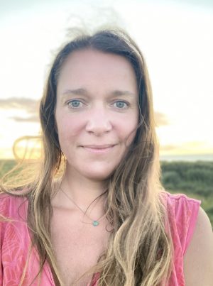 Eine Person mit langen Haaren und einem rosa Oberteil steht im Freien vor einer grünen Kulisse und einem hellen Himmel. Sie hat einen ruhigen Gesichtsausdruck, trägt eine Halskette mit einem kleinen Anhänger und ihre Präsenz scheint leise zu sagen: „Über mich.“